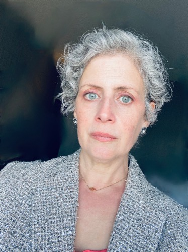 A person with short gray hair and blue eyes, perhaps named Preston or Rebecca, looks directly at the camera. They wear a textured gray jacket and small earrings, standing against a dark background.
