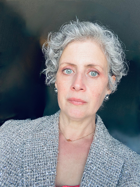 A person with short gray curly hair and pearl earrings, reminiscent of Rebecca Prestons iconic style, wears a textured blazer and looks directly at the camera. The dark background accentuates their striking features and sophisticated attire.