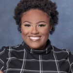 Smiling Kyra Robinson with short curly hair wears a black shirt adorned with white grid patterns, set against a serene blue background.