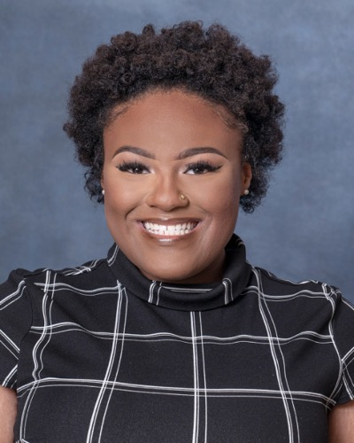 Smiling Kyra Robinson with short curly hair wears a black shirt adorned with white grid patterns, set against a serene blue background.