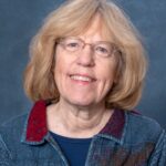 Elizabeth Sale, a woman with shoulder-length blonde hair, is wearing glasses and a blue patterned jacket over a dark shirt. She smiles warmly against a plain gray-blue background.