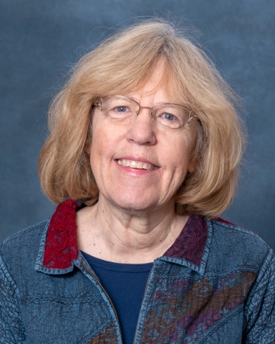 Elizabeth Sale, a woman with shoulder-length blonde hair, is wearing glasses and a blue patterned jacket over a dark shirt. She smiles warmly against a plain gray-blue background.