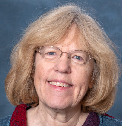 An older person with shoulder-length blond hair and glasses, reminiscent of Elizabeth, smiles against a gray-blue background. They are wearing a sweater with a multicolored collar.