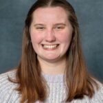 Hannah Schleicher, a woman with long brown hair, smiles warmly while wearing a light gray knitted sweater against a plain dark blue background.