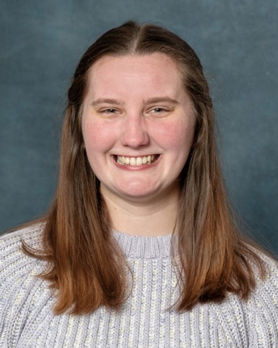Hannah Schleicher, a woman with long brown hair, smiles warmly while wearing a light gray knitted sweater against a plain dark blue background.