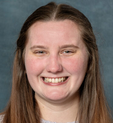 Hannah, with her long brown hair, smiles warmly against a blue-gray background.