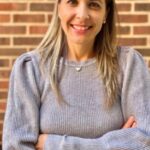 Esmarie, with her long blonde hair and gray sweater, stands confidently against the brick wall, arms crossed and smiling warmly at the camera.