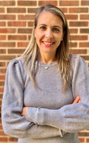 Esmarie, with her long blonde hair and gray sweater, stands confidently against the brick wall, arms crossed and smiling warmly at the camera.