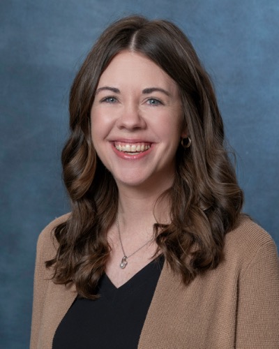 Rachel Taube, with her long brown hair, smiles warmly in a black top and brown cardigan against a gray-blue background.