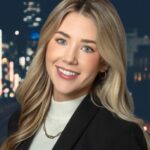 Lindsey, a smiling woman with long blonde hair, dons a black blazer and white turtleneck as she stands against a blurred cityscape at night.