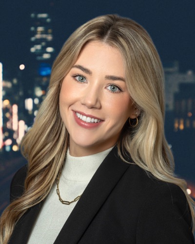 Lindsey, a smiling woman with long blonde hair, dons a black blazer and white turtleneck as she stands against a blurred cityscape at night.