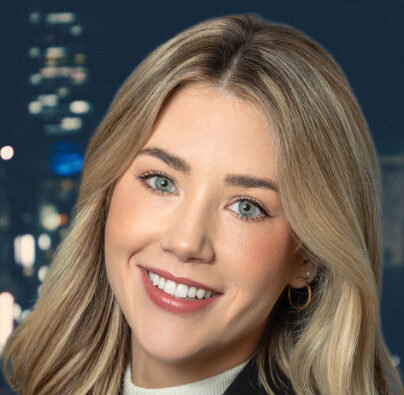 With long blonde hair, Lindsey smiles warmly at the camera. She dons a dark blazer with a light-colored top, set against a blurred nighttime cityscape illuminated by twinkling lights.