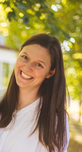 Claire, a woman with long brown hair, is smiling warmly at the camera. She is wearing a white blouse while standing outdoors against a backdrop of blurred trees, suggesting its a sunny day.