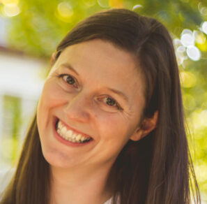 Claire, with her long brown hair cascading down her shoulders, stands outdoors with a bright smile. The blurred green and yellow foliage in the background hints at a sunny day, creating a serene ambiance around Claires presence.