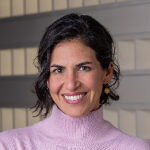 Rachel, with short black hair, beams in a pink turtleneck and gold earrings, set against a striking geometric patterned background.