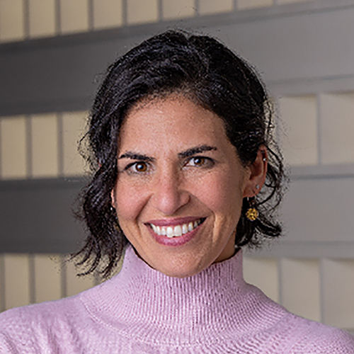 Rachel, with short black hair, beams in a pink turtleneck and gold earrings, set against a striking geometric patterned background.