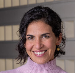 Rachel, with short dark hair, wearing a light pink turtleneck and gold earrings, smiles at the camera. The background features a patterned wall with beige and gray tones, capturing a warm and inviting essence synonymous with Winograds aesthetic.