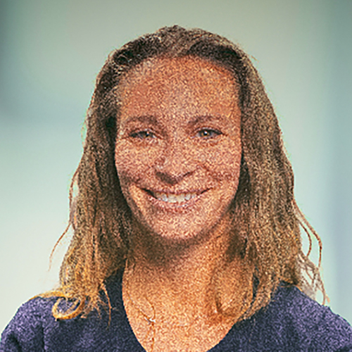 Smiling Jodi, with wavy brown hair, dons a dark top against a light-colored background.