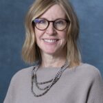 Amy, with shoulder-length blonde hair and glasses, smiles at the camera. She wears a beige top and a layered beaded necklace against a blue background.
