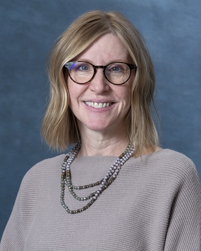 Amy, with shoulder-length blonde hair and glasses, smiles at the camera. She wears a beige top and a layered beaded necklace against a blue background.