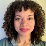 Elizabeth, with her curly hair and denim shirt, smiles warmly at the camera. Against a plain, light-colored wall, she exudes effortless charm.