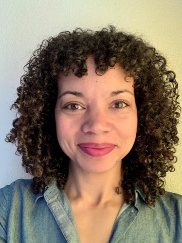 Elizabeth, with her curly hair and denim shirt, smiles warmly at the camera. Against a plain, light-colored wall, she exudes effortless charm.