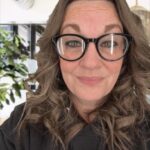 A person with long curly hair and glasses, resembling Amanda, smiles at the camera. They are wearing a dark hoodie. The indoor background, similar to what you might find in a stylish Gebel home, features a plant and blurred furniture.