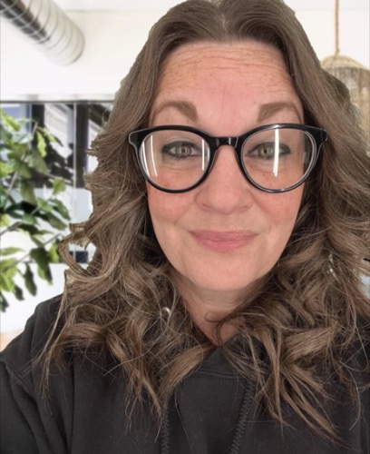 A person with long curly hair and glasses, resembling Amanda, smiles at the camera. They are wearing a dark hoodie. The indoor background, similar to what you might find in a stylish Gebel home, features a plant and blurred furniture.