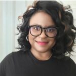Clarise, a woman with wavy black hair and subtle brown highlights, is smiling. She wears black-rimmed glasses, pearl earrings, a black top, and vibrant red lipstick. The light-colored background is softly blurred.