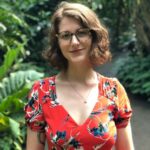 Lindsey Pawlowski, with shoulder-length brown hair and glasses, stands smiling in a lush garden. She wears a red floral dress with a V-neck. The background features greenery, including large leaves and plants along a pathway.