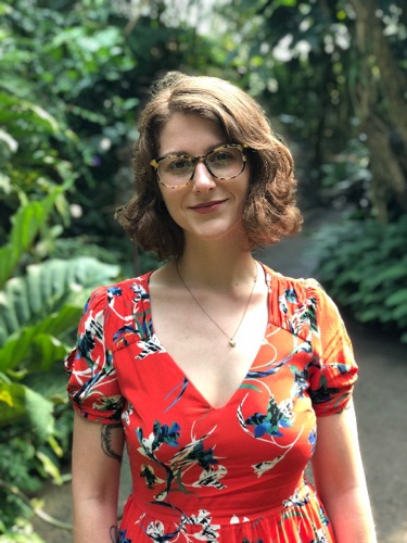 Lindsey Pawlowski, with shoulder-length brown hair and glasses, stands smiling in a lush garden. She wears a red floral dress with a V-neck. The background features greenery, including large leaves and plants along a pathway.