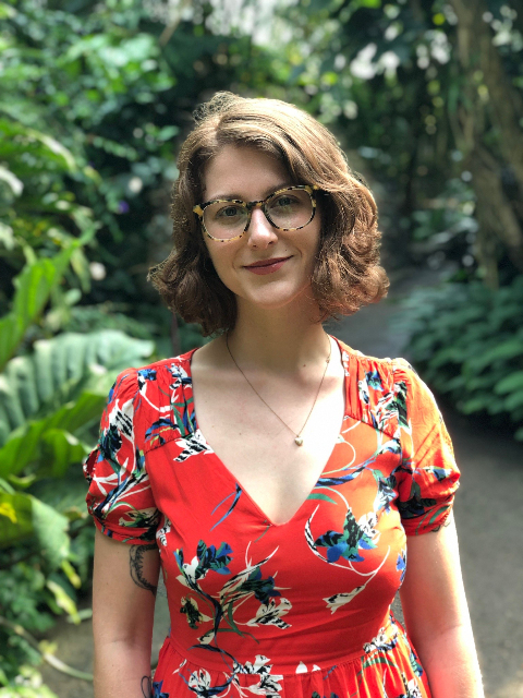 Lindsey Pawlowski, with shoulder-length brown hair and glasses, stands in a lush garden wearing a red floral dress and smiling slightly. The background is filled with vibrant green leaves and plants.