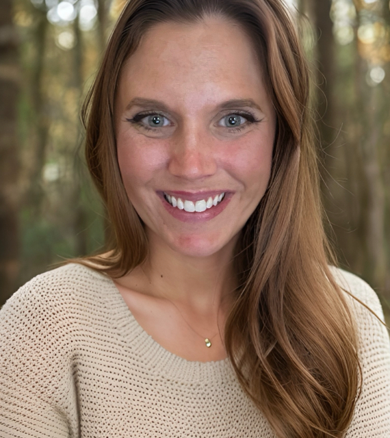 A smiling woman, Sarah Phillips, with long brown hair wearing a beige sweater is standing outdoors with trees in the background.
