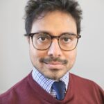 Siddiqui Saad, a man with short dark hair and glasses, wears a burgundy sweater over a blue checkered shirt and navy tie. He looks directly at the camera with a neutral expression against a plain background.