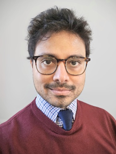 Siddiqui Saad, a man with short dark hair and glasses, wears a burgundy sweater over a blue checkered shirt and navy tie. He looks directly at the camera with a neutral expression against a plain background.