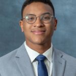 A person wearing glasses and a gray suit with a white polka-dot shirt and blue tie smiles against a blue background, embodying the polished style of Joshua Wilson.