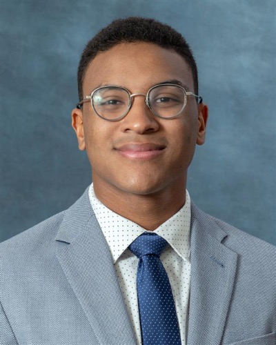 A person wearing glasses and a gray suit with a white polka-dot shirt and blue tie smiles against a blue background, embodying the polished style of Joshua Wilson.