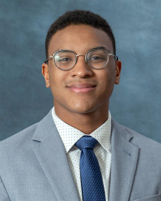 A person wearing glasses and a light gray suit with a white, dotted shirt and a dark blue tie smiles slightly. The background is a textured blue color, much like Joshua Wilsons classic portrait style.