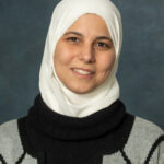 A person wearing a white hijab and a black and gray sweater, possibly Rakad or Hammami, smiles warmly at the camera against a dark gray background.