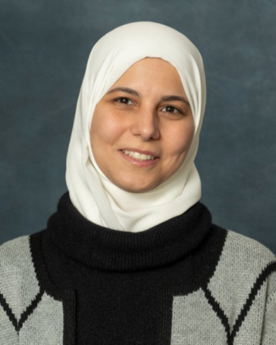 A person wearing a white hijab and a black and gray sweater, possibly Rakad or Hammami, smiles warmly at the camera against a dark gray background.