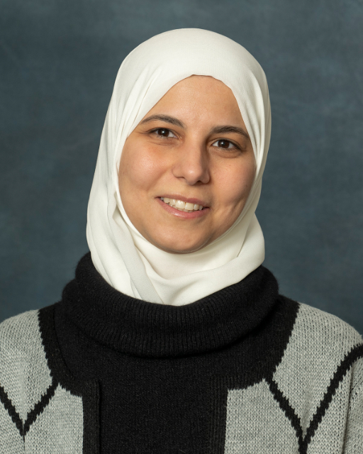 A person wearing a white headscarf and a black and gray Rakad sweater smiles against a gray background.