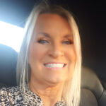 Karen, with her long blonde hair, sits in a Bartoni car as sunlight filters through the window, highlighting her cheerful face. She wears a patterned blouse and a delicate necklace, perfectly complementing the serene moment.