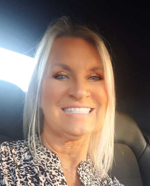 Karen, with her long blonde hair, sits in a Bartoni car as sunlight filters through the window, highlighting her cheerful face. She wears a patterned blouse and a delicate necklace, perfectly complementing the serene moment.