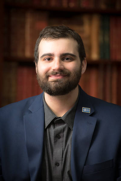A person with a beard and short hair, possibly Jake Agliata, is wearing a dark blue blazer and gray shirt, smiling at the camera. The background consists of shelves filled with books stacked closely together.