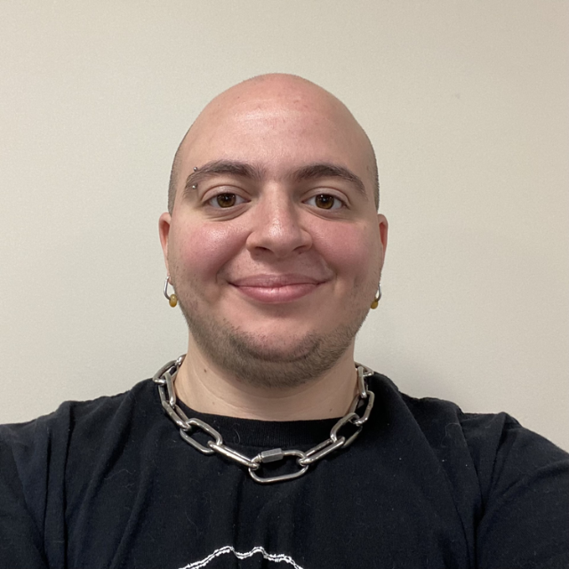 James Lituchy, sporting a shaved head, black t-shirt, chain necklace, and earrings, smiles confidently at the camera against a plain background.