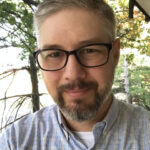 Isaac Sandidge, a man with short gray hair, a beard, and glasses, wearing a light blue checkered shirt, smiles slightly while standing outdoors in front of trees and greenery.