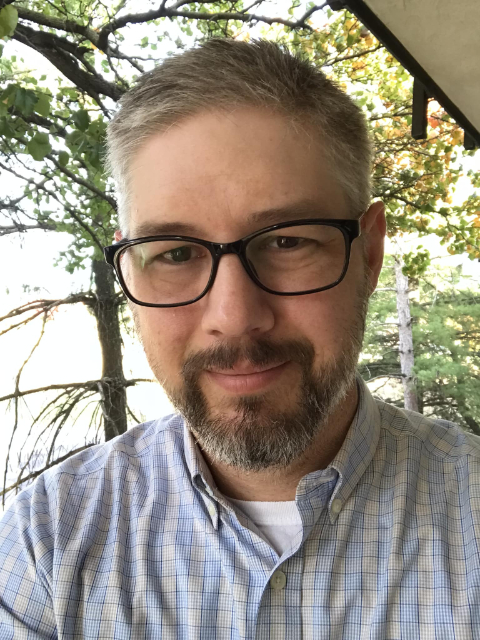 Isaac Sandidge, a man with short gray hair, a beard, and glasses, wearing a light blue checkered shirt, smiles slightly while standing outdoors in front of trees and greenery.