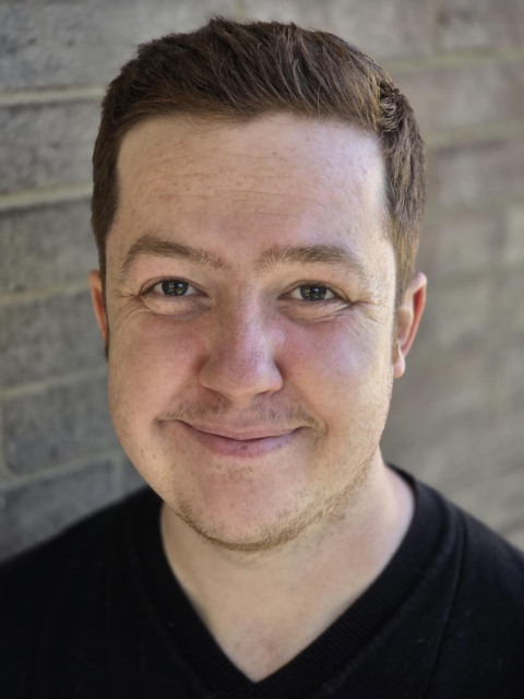 A person with short brown hair and fair skin is smiling warmly at the camera, standing in front of a brick wall and wearing a black V-neck top.