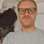 Gee Everett, a man with light hair, a beard, and glasses, wears a grey t-shirt as he stands indoors. Behind him, a backpack hangs on the wall beside exposed pipes and a whiteboard.