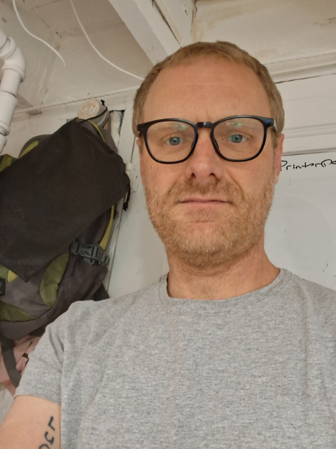 Gee Everett, a man with light hair, a beard, and glasses, wears a grey t-shirt as he stands indoors. Behind him, a backpack hangs on the wall beside exposed pipes and a whiteboard.